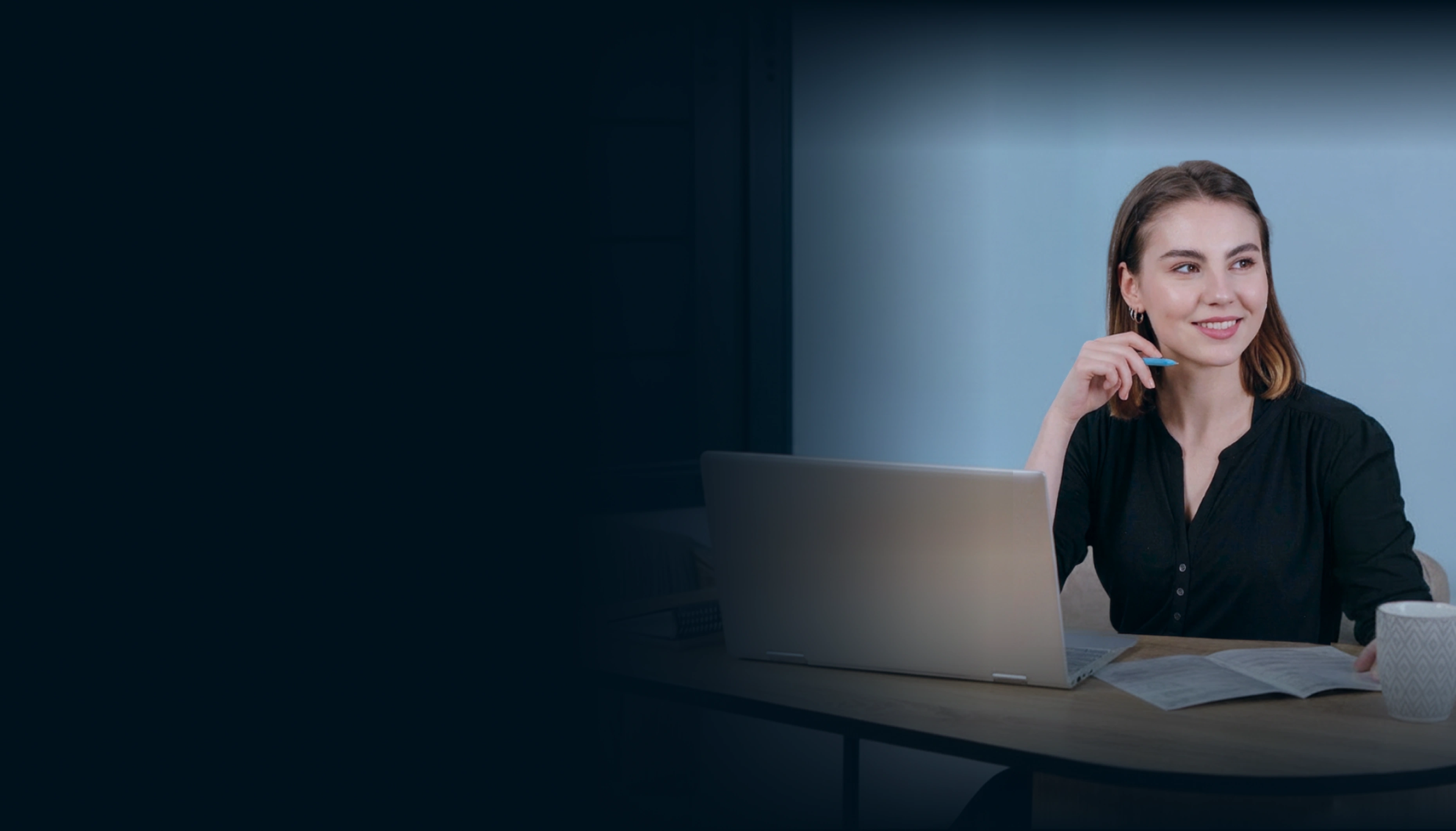 A woman works at a desktop computer in a office, thinking and smiling.