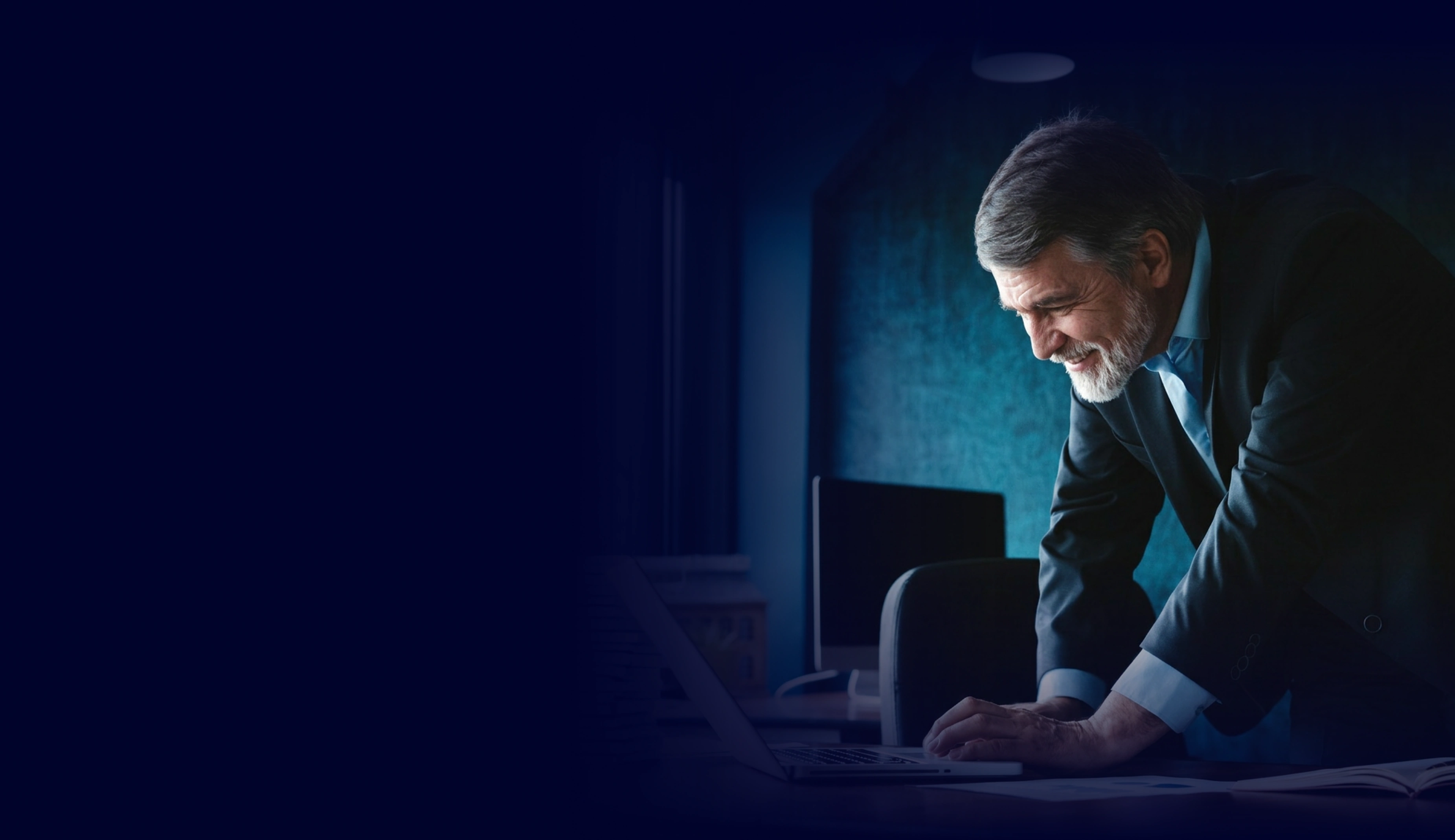 Older man in a suit working on a laptop at a desk in a dimly lit office.